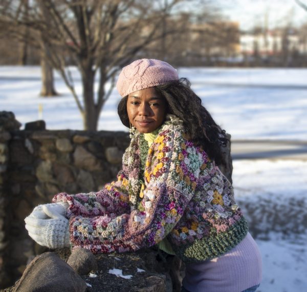 girl standing outside wearing a pink, green, yellow and cream granny square sweater. The sweater has a button closure