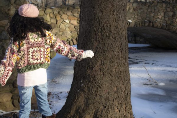 girl standing outside wearing a pink, green, yellow and cream granny square sweater. The sweater has a button closure