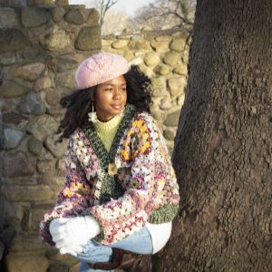 girl standing outside wearing a pink, green, yellow and cream granny square sweater. The sweater has a button closure