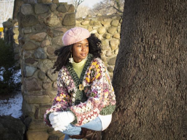 girl standing outside wearing a pink, green, yellow and cream granny square sweater. The sweater has a button closure