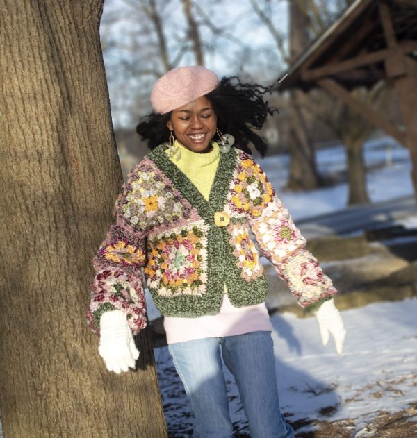 girl standing outside wearing a pink, green, yellow and cream granny square sweater. The sweater has a button closure