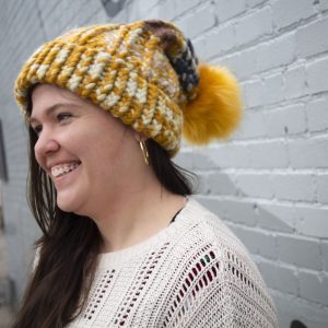 girl in handknit floppy golden yellow and navy blue toboggan. The toboggan has a pom pom at the end