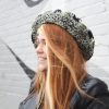 girl standing against a gray way wearing a hand crocheted olive green, black and gray beret or tam with black pom pom