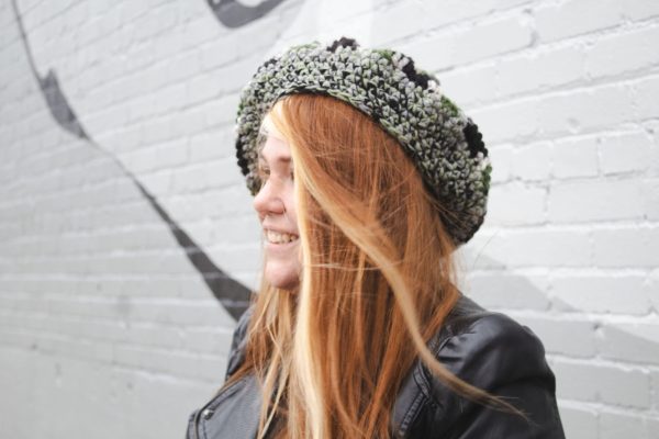 girl standing against a gray way wearing a hand crocheted olive green, black and gray beret or tam with black pom pom