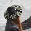 girl standing against a gray way wearing a hand crocheted olive green, black and gray beret or tam with black pom pom
