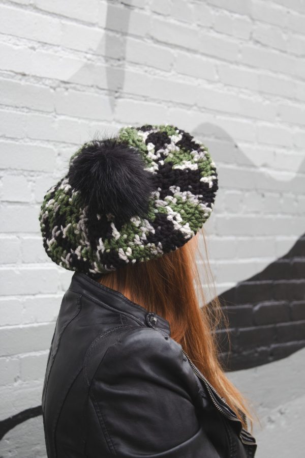 girl standing against a gray way wearing a hand crocheted olive green, black and gray beret or tam with black pom pom