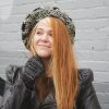 girl standing against a gray way wearing a hand crocheted olive green, black and gray beret or tam with black pom pom