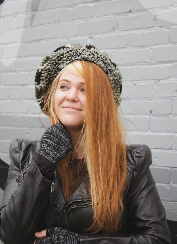 girl standing against a gray way wearing a hand crocheted olive green, black and gray beret or tam with black pom pom