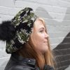 girl standing against a gray way wearing a hand crocheted olive green, black and gray beret or tam with black pom pom