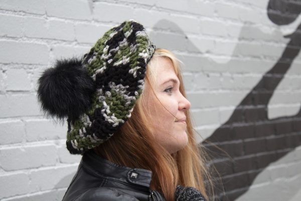 girl standing against a gray way wearing a hand crocheted olive green, black and gray beret or tam with black pom pom