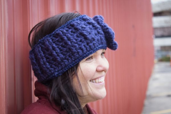 girl wearing a navy blue handmade crocheted headband or ear warmer. The ear warmer has a hand crocheted flower with a brown button center