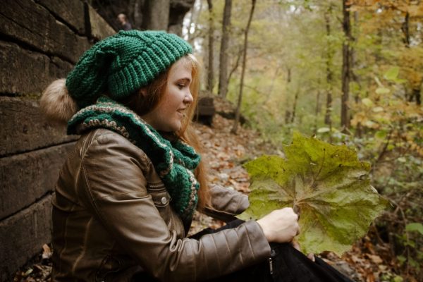 Girl sitting outside wearing a vibrant green floppy toboggan and infinity scarf matching set. The toboggan and scarf are handmade by MiasWorkshop. The hat is hand knit. The scarf is crocheted. The toboggan has a tan pom pom attached. The scarf has a matching tan stripe running through it.