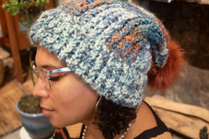 girl wearing a handmade knit slouchy toboggan. The toboggan is blue, green, cram, and orange, and it has a matching orange pom pom