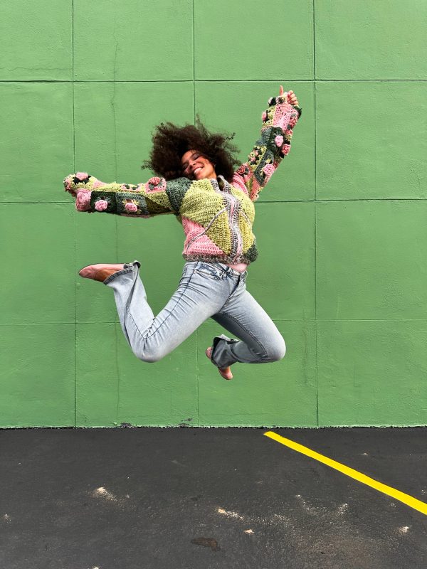 girl with curly hair against a green background wearing a pink and green granny square sweater with roses on the sleeves. The sweater zips in the front and has pockets