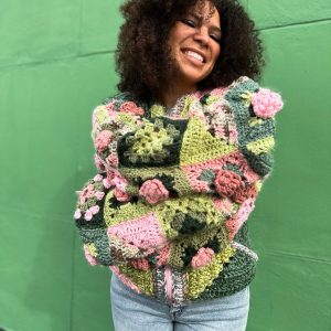 girl with curly hair against a green background wearing a pink and green granny square sweater with roses on the sleeves. The sweater zips in the front and has pockets