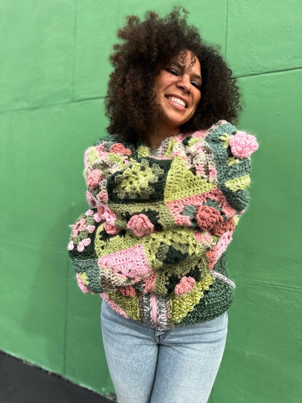 girl with curly hair against a green background wearing a pink and green granny square sweater with roses on the sleeves. The sweater zips in the front and has pockets