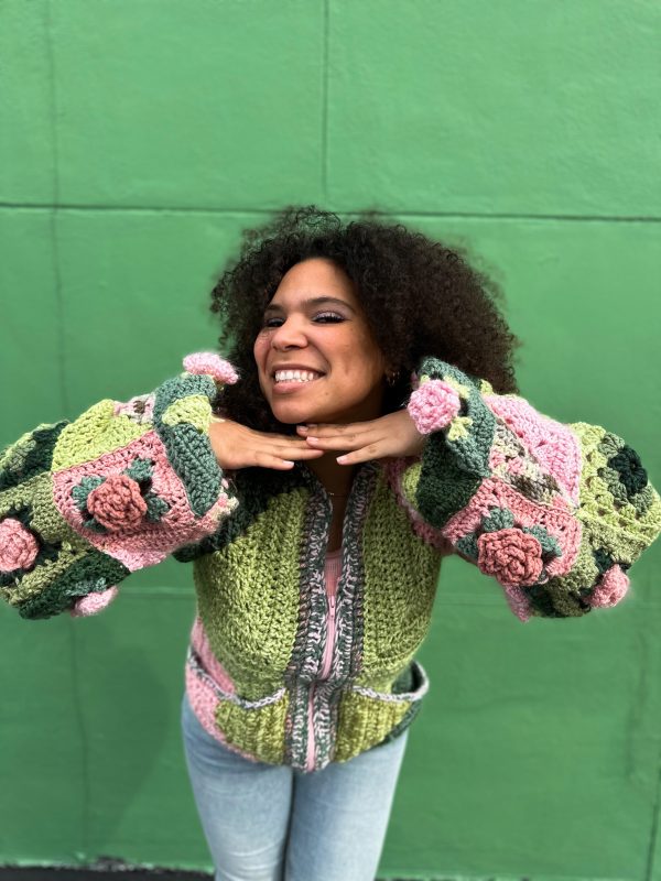 girl with curly hair against a green background wearing a pink and green granny square sweater with roses on the sleeves. The sweater zips in the front and has pockets