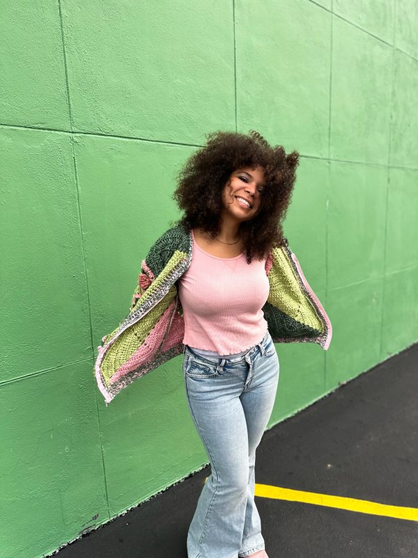 girl with curly hair against a green background wearing a pink and green granny square sweater with roses on the sleeves. The sweater zips in the front and has pockets