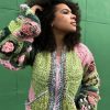 girl with curly hair against a green background wearing a pink and green granny square sweater with roses on the sleeves. The sweater zips in the front and has pockets