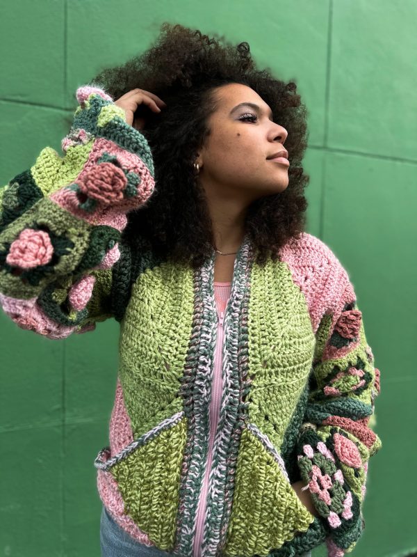 girl with curly hair against a green background wearing a pink and green granny square sweater with roses on the sleeves. The sweater zips in the front and has pockets