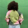 girl with curly hair against a green background wearing a pink and green granny square sweater with roses on the sleeves. The sweater zips in the front and has pockets