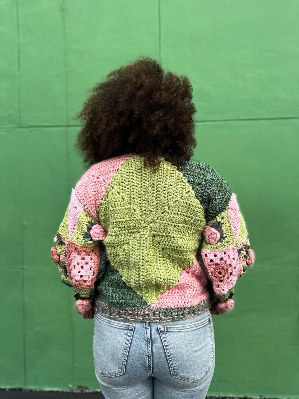 girl with curly hair against a green background wearing a pink and green granny square sweater with roses on the sleeves. The sweater zips in the front and has pockets