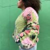 girl with curly hair against a green background wearing a pink and green granny square sweater with roses on the sleeves. The sweater zips in the front and has pockets