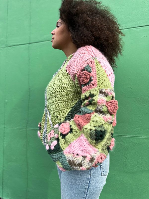 girl with curly hair against a green background wearing a pink and green granny square sweater with roses on the sleeves. The sweater zips in the front and has pockets