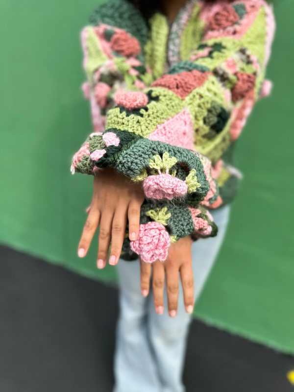 girl with curly hair against a green background wearing a pink and green granny square sweater with roses on the sleeves. The sweater zips in the front and has pockets