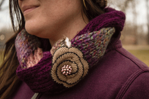 Girl standing outside wearing a braided cowl scarf with multiple colors of violet, brown and cream. The cowl scarf also has a light brown fabric flower accent with beaded center