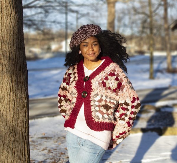 a girl standing outside in a pink, dark red, dark brown, and cream granny square sweater. The sweater buttons up the front. MiasWorkshop