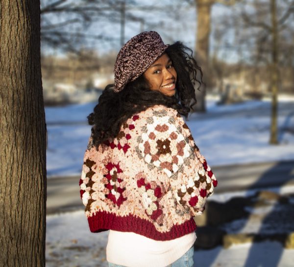 a girl standing outside in a pink, dark red, dark brown, and cream granny square sweater. The sweater buttons up the front. MiasWorkshop