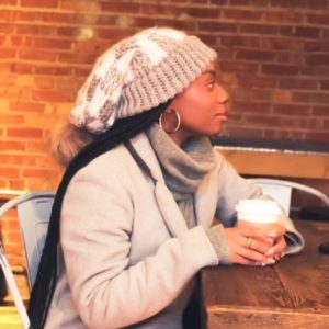 girl in a coffee shop wearing a handmade, knit, slouchy, fuzzy, cream and gray checkered toboggan with a gray pom pom attached